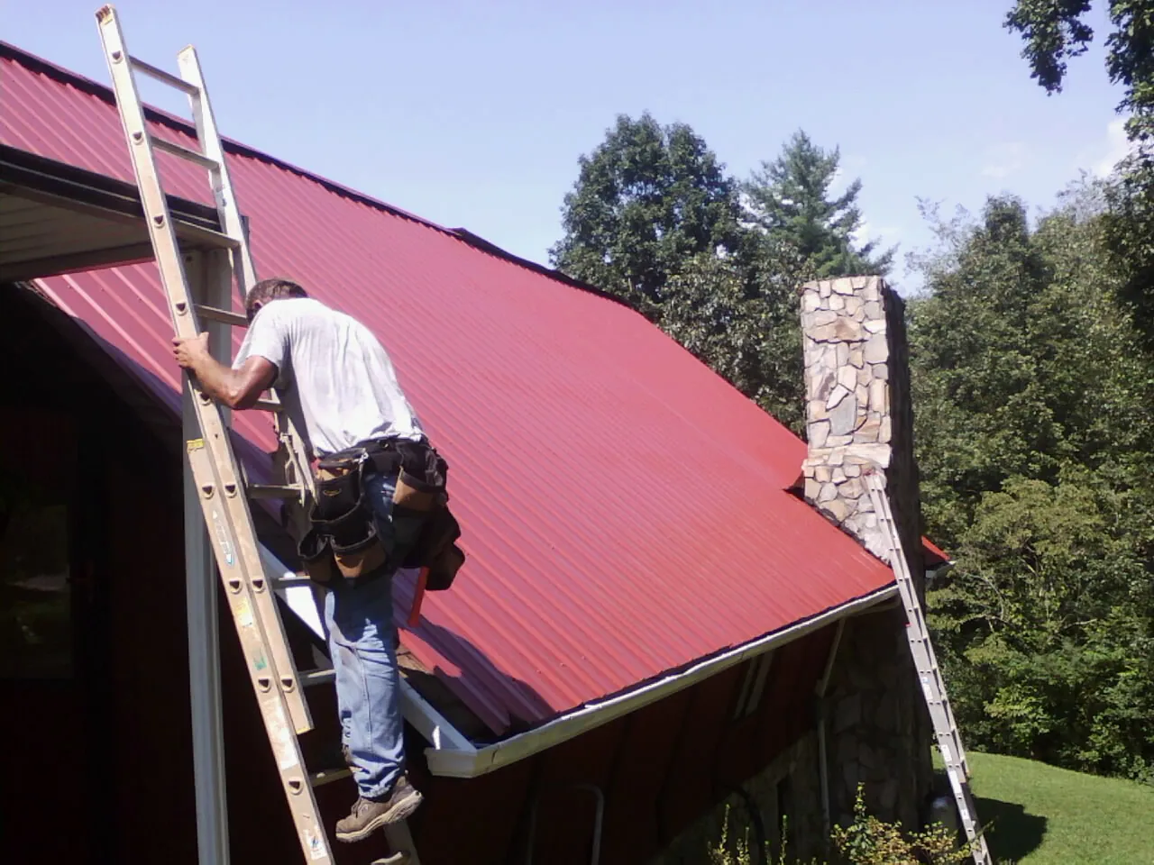 Roofer on a ladder beside a red metal roof and stone chimney — Soffit & Fascia Repair by Roofing Brothers in Columbia